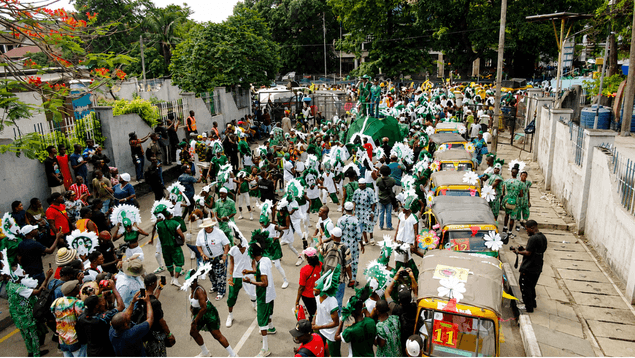 Green and white carnival procession beside flower-decorated yellow kekes